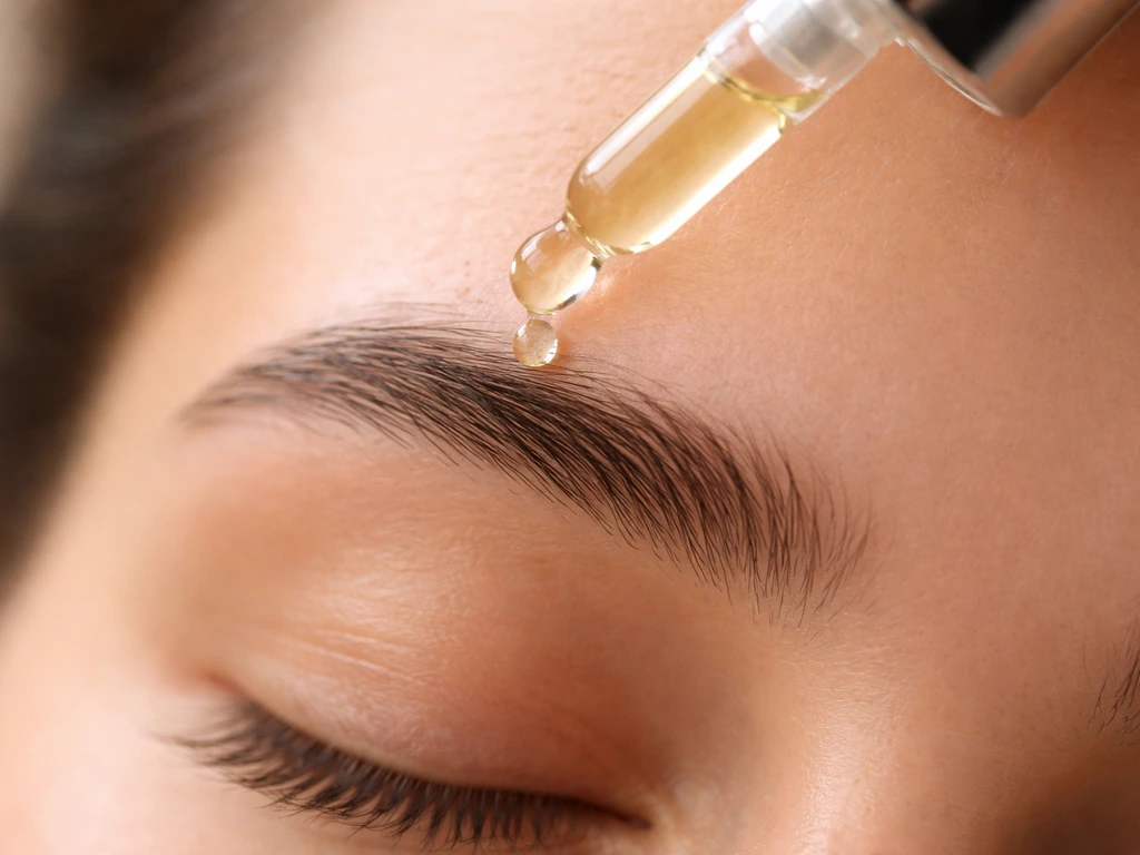 A dropper applies clear vitamin E oil along an eyebrow line in a close-up, natural-light photo.